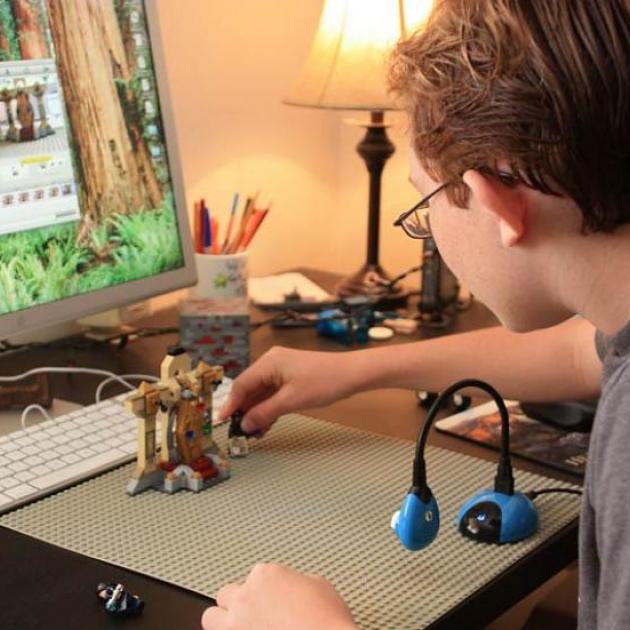 A boy creates a stop motion movie with a Hue Animation Kit. The boy sits at a desk, with the desktop screen in the background showing the Hue animation software - a split screen for showing the last shot and new positioning of the character. The boy adjusts one of his props. A blue Hue animation camera - a small webcam with a flexible neck that can be positioned anyway - sits on the desk capturing the image. 