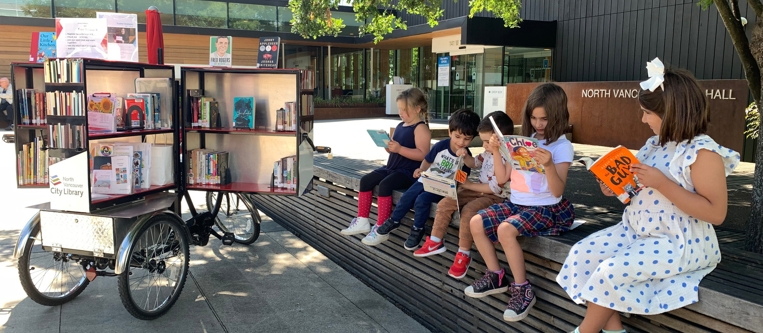 A tricycle with a large unit on the back that is open and full of books, children sitting reading