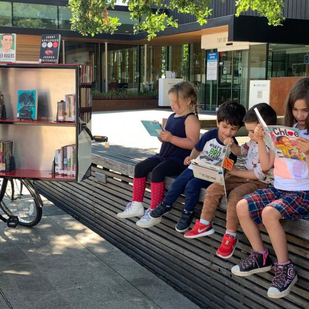Book Bike A tricycle with a large unit on the back that is open and full of books, children sitting reading
