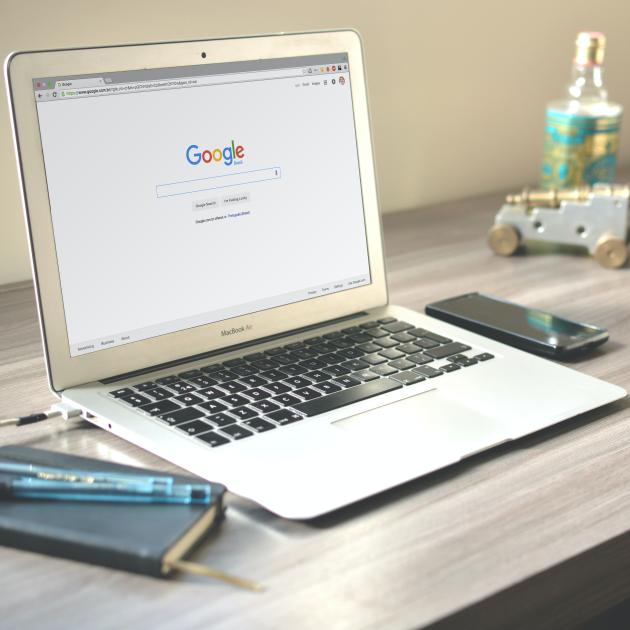 Google Macbook laptop sits open on a desk, with a browser open on a Google search bar. The laptop sits on a light wooden desk. There is a small black notebook on the left hand side with two pens resting on top. On the left there is a black cell phone and some out of focus desk decorations.