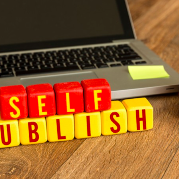 Self Publish Stackable letter blocks sit on a table and read "Self Publish". The "Self" blocks are red with yellow letters. The "Publish" blocks are yellow with red letters. A Macbook laptop sits behind the blocks. A orange mug with saucer sits to the side with coffee in it.