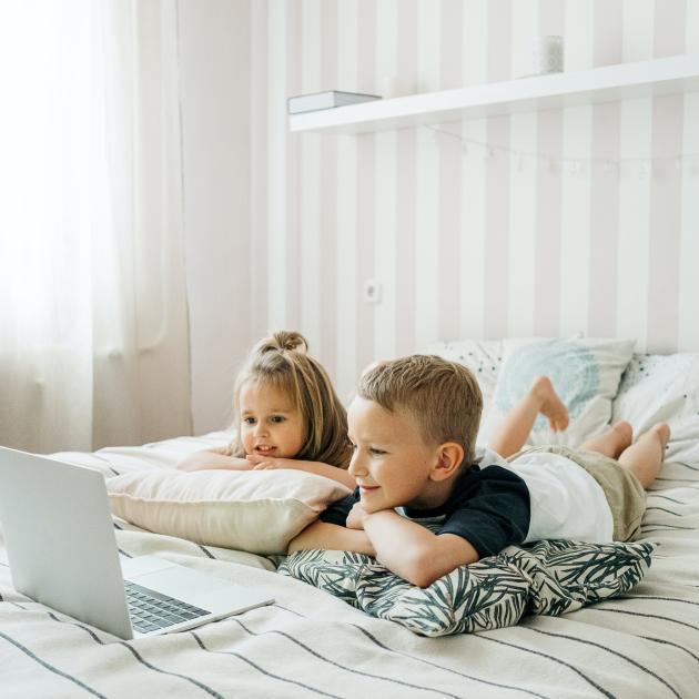 A girl and a boy lying on a bed watching something on a laptop.