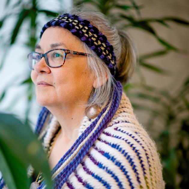 Photo portrait of Kung Jaadee with regalia shawl and headband, wearing glasses, looking to the left with plants in background.