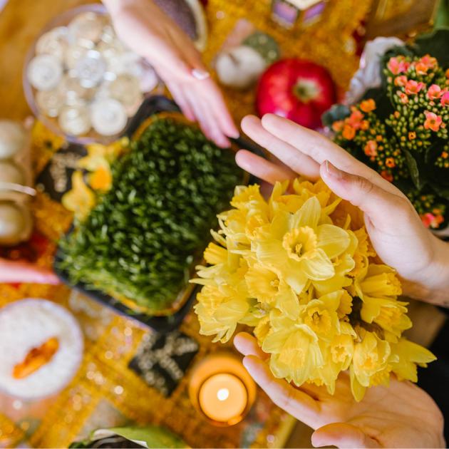 Nowruz haft-sin table with \hHands holding bunch of yellow daffodils, green shoots, red apple and candles