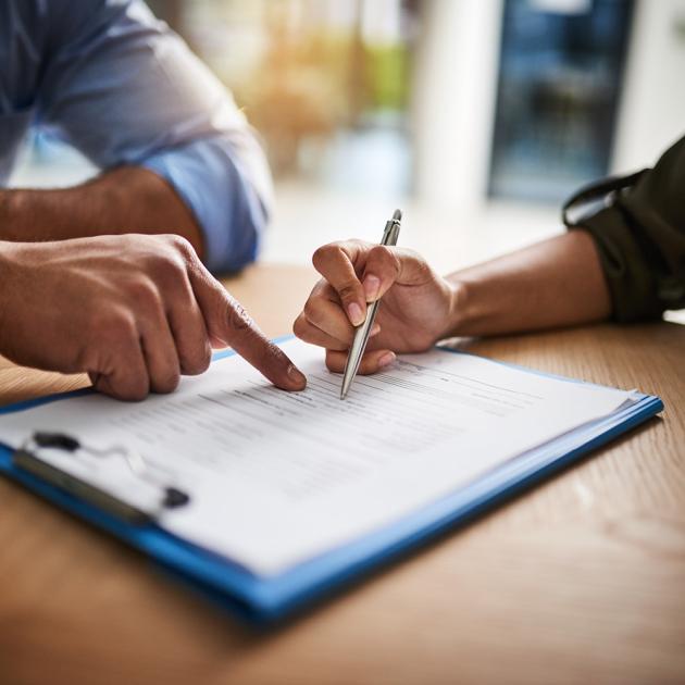 two people's hands, one pointing at clipboard holding paperwork, one ready to sign with a pen