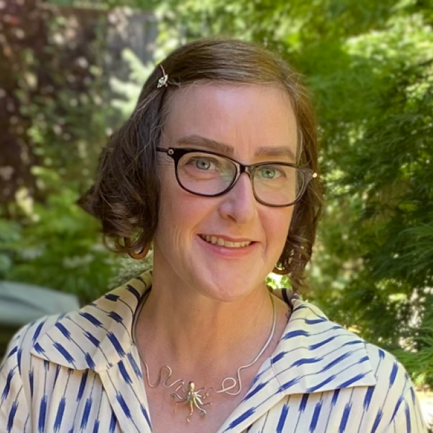 Erin MacNair, local author A woman with light brown hair in the curly bob and glasses, faces the camera against a green background.