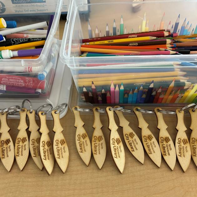 Paddle keychain craft Small regalia paddles with NVCL logo engraved on them lined in front of pencil crayons and markers.