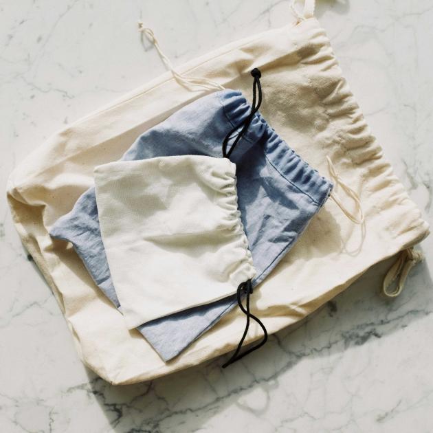 The drawstring bags sit on a marble surface. Three drawstring bags sit piled together on a marble surface. The largest and smallest bags are cream-coloured and the middle-size bag is light blue.