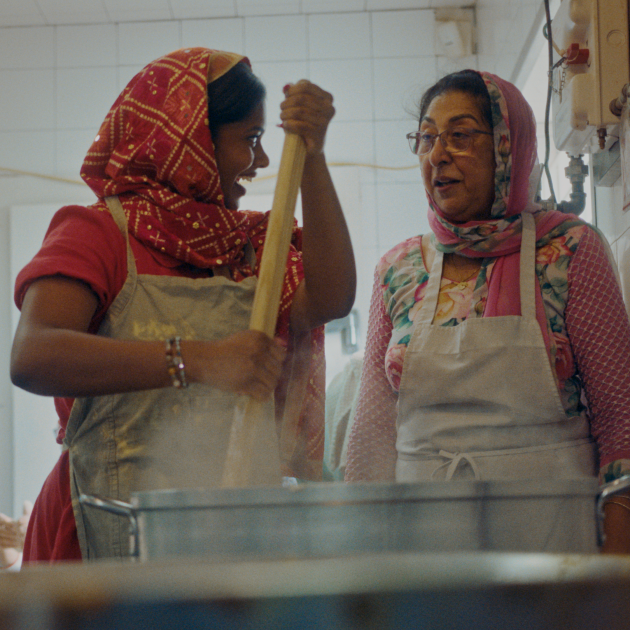 Not Your Butter Chicken Two South Asian women wear aprons in a kitchen, while one stirs a pot with a big spoon