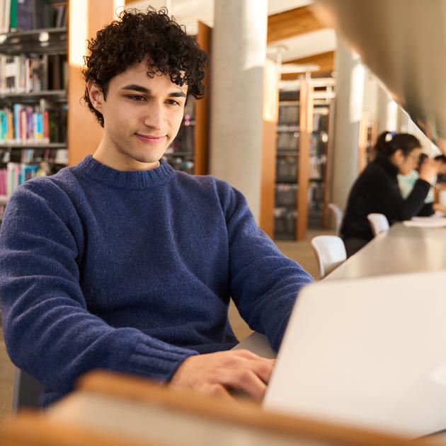Writing for university applications Young adult in library sitting at desk with laptop open, looking at the screen