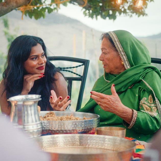 Two woman sit at outdoor dining table, talking, with large dishes of food in front of them