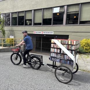a man on a bicycle towing a mini book library on wheels