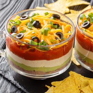 Glass with layers of bean dip in red, white and green colours with cheese on top, tortilla chips in foreground