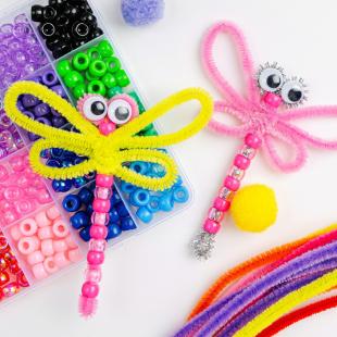  A yellow, pipe cleaner dragonfly with light and dark colored pink beads and googly eyes.  A pink dragonfly with pink pipe cleaners and pink beads. Box of colored beads and colored pipe cleaners.
